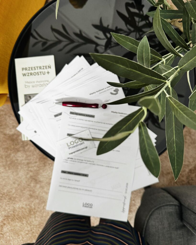 Materiały warsztatowe Przestrzeni Wzrostu i LOGO PRZESTRZEŃ na stoliku obok gałązki oliwnej